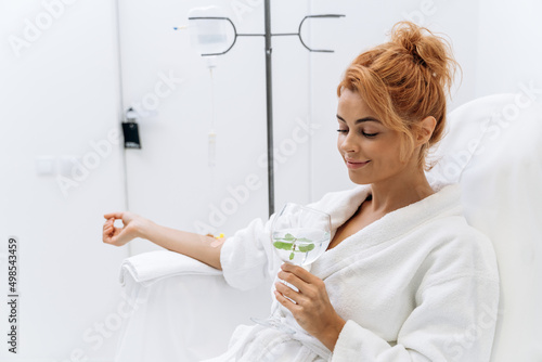 Woman in white bathrobe sitting in armchair and receiving IV infusion