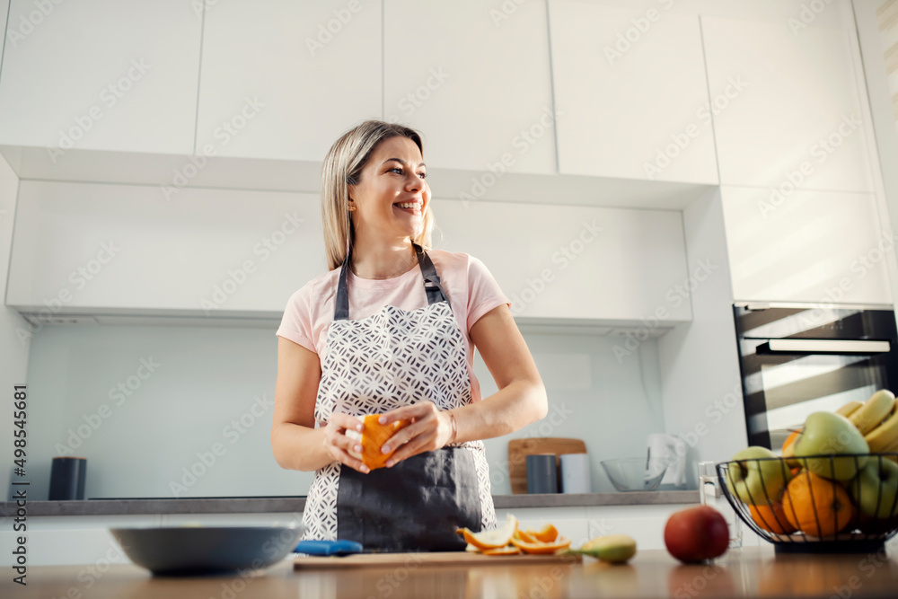 A smiling woman in kitchen peeling orange and preparing fruit salad.