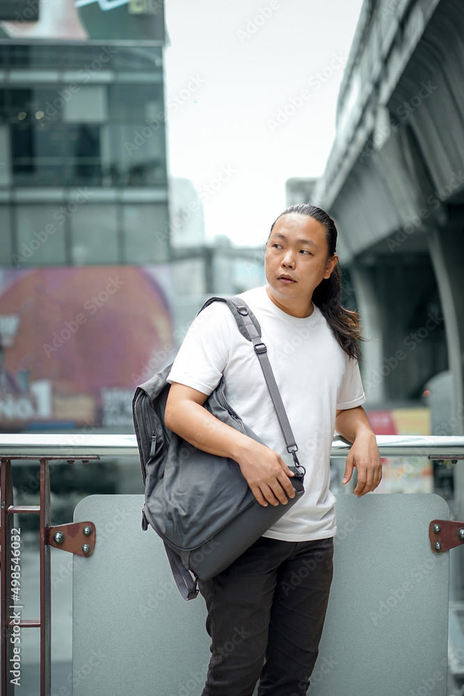 Long Hair Thai, Chinese man is posing in front of the terrace barricade with gray back on the shoulder at outdoor building environment Long Hair Thai, Chinese man is posing in front of the terrace barricade with gray back on the shoulder at outdoor building environment