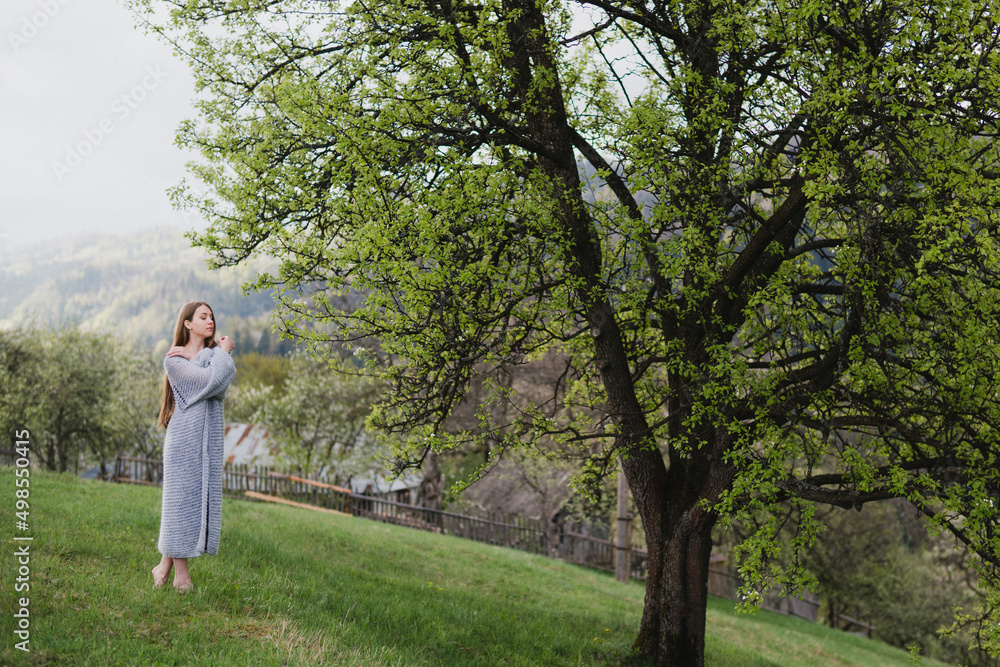 Young pretty emotional caucasian woman with long hair wearing natural makeup in romantic casual clothes walking among spring mountains near the beautiful fresh blooming tree in the countryside.