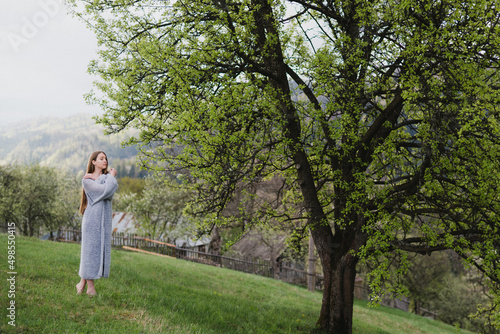 Young pretty emotional caucasian woman with long hair wearing natural makeup in romantic casual clothes walking among spring mountains near the beautiful fresh blooming tree in the countryside.