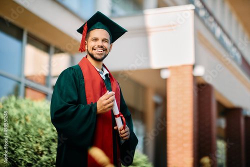 Happy university student in graduation gown holding his diploma and looking at camera. photo