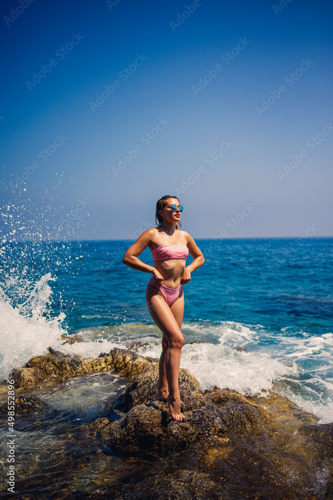 Beautiful young woman in a swimsuit on a rocky beach on a sunny day against the backdrop of waves. Vacation in the summer season. Selective focus Beautiful young woman in a swimsuit on a rocky beach on a sunny day against the backdrop of waves. Vacation in the summer season. Selective focus