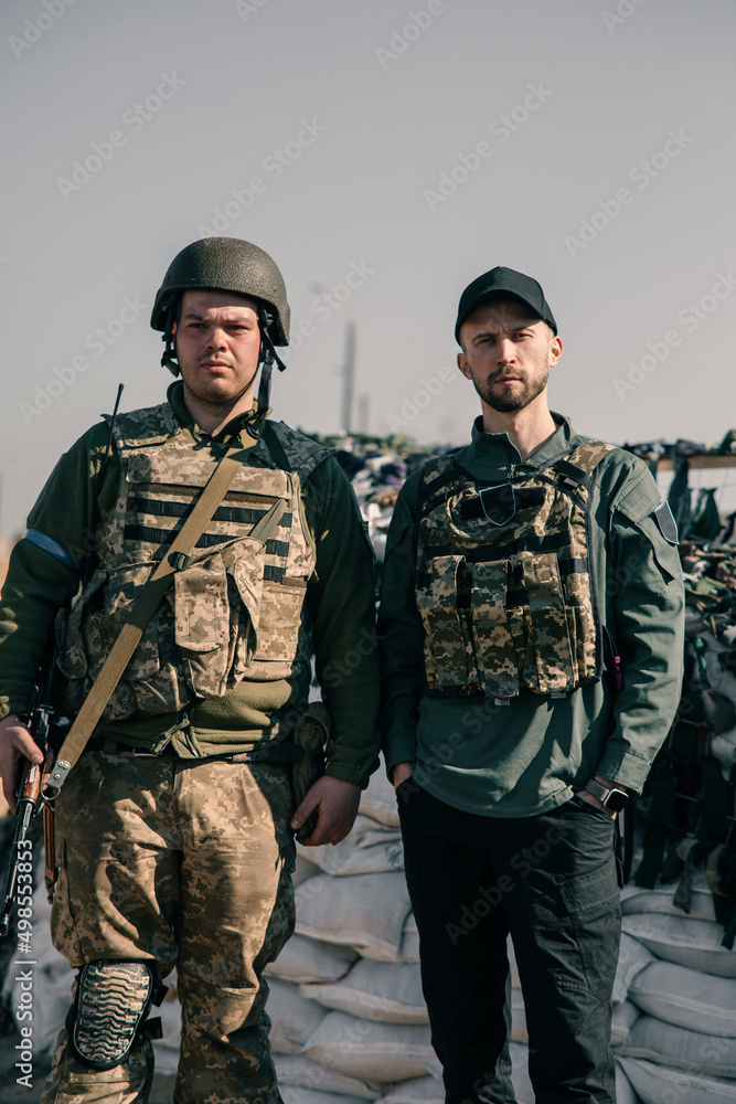 Ukrainian territorial defense warrior stands near volunteer on checkpoint in body armor with automatic gun. Ukrainian territorial defense warrior stands near volunteer on checkpoint in body armor with automatic gun.