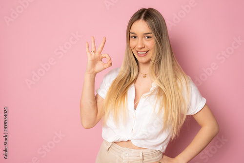 Happy smiling young woman showing okay sign standing isolated over pink background.