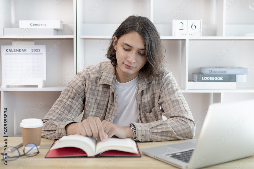 Young male student reading a book in a private office, Long distance education, Textbooks from books, Find out more, In the office there is a computer or laptop placed on the table, Study. Young male student reading a book in a private office, Long distance education, Textbooks from books, Find out more, In the office there is a computer or laptop placed on the table, Study.