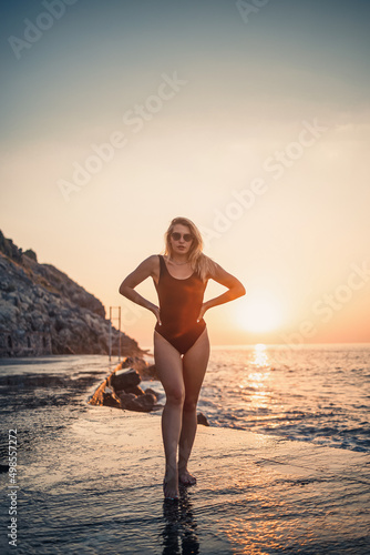 Beautiful young woman in a black swimsuit alone on the beach by the sea at sunset. An adult slender girl resting on the ocean shore at sunset. Summer vacation concept. Selective focus