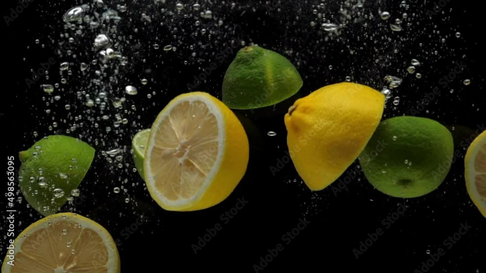 Slow motion close-up yellow lemon and green lime halves falling into transparent water on black background. Fresh fruits and berries splashing in aquarium. Citrus, healthy food, diet, air bubbles