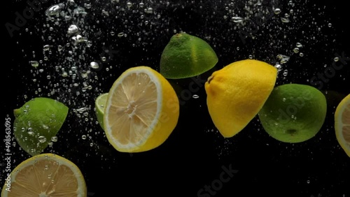 Slow motion close-up yellow lemon and green lime halves falling into transparent water on black background. Fresh fruits and berries splashing in aquarium. Citrus, healthy food, diet, air bubbles