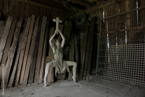 A demon like figure dressed in rags holds a crucifix up high in her hands. The horror figure wears a floral thorny crown on her head. 