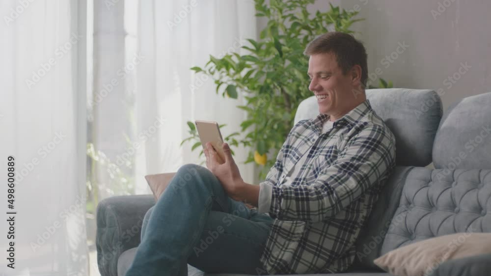 Cheerful man usinng tablet and chatting with friends sitting on sofa in living room, modern style room with loft interior Cheerful man usinng tablet and chatting with friends sitting on sofa in living room, modern style room with loft interior