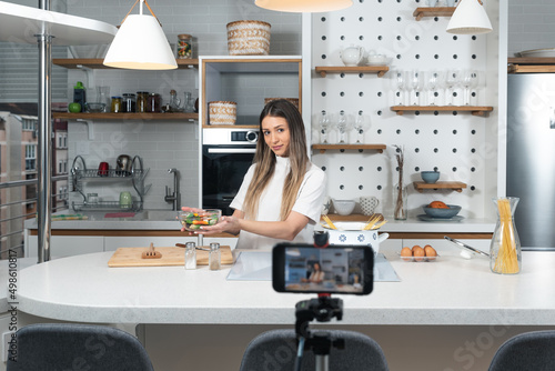 Fotografie Young vlogger woman vlogging about cooking at her apartment domestic kitchen cook and making video vlog with smartphone as online content creator for internet