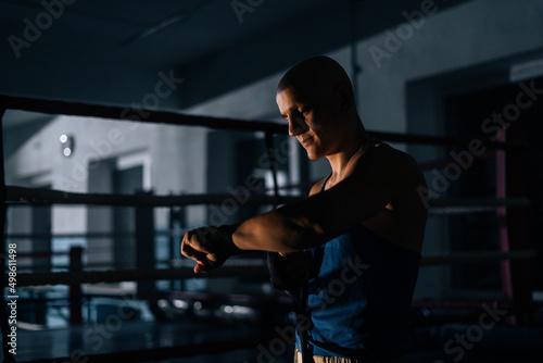 Side view of serious bald boxer wrapping defense boxing tape around wrist before fight in sport club with dark interior on background of ring. Serious fighter getting ready for fight in fitness center