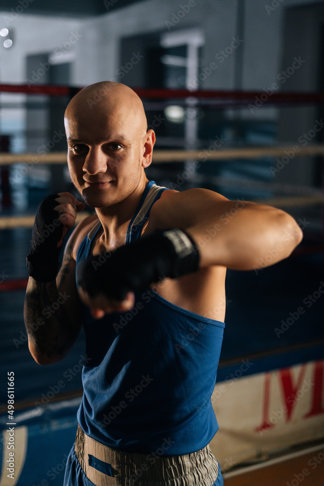 Vertical shot of bald aggressive boxer male wearing bandages punching air to camera in sport club with dark interior, on background of ring. Professional fighter fighting shadow looking at camera. Vertical shot of bald aggressive boxer male wearing bandages punching air to camera in sport club with dark interior, on background of ring. Professional fighter fighting shadow looking at camera.
