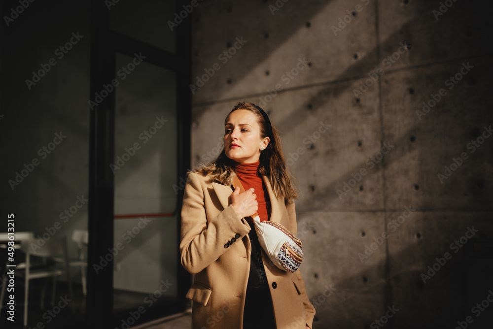 young woman on the background of a building on which the rays of sunlight fall, a woman holds her hand on a waist bag young woman on the background of a building on which the rays of sunlight fall, a woman holds her hand on a waist bag