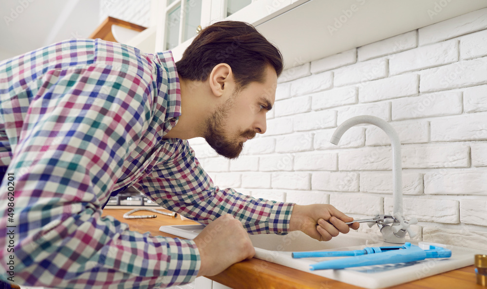 Plumber at work. Young repairman fixing the tap at home. Serious handsome man bending over the sink and using his tools to repair or install a modern mixer faucet in the kitchen