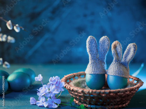 Easter eggs with crocheted hats with bunny ears in a wicker basket. Blue background copy space