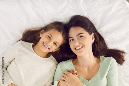 Happy mother together with child looking at camera and smiling while resting on soft white pillow on warm bed at home. Family, sleep, health and beauty concept. High angle shot, top view, from above