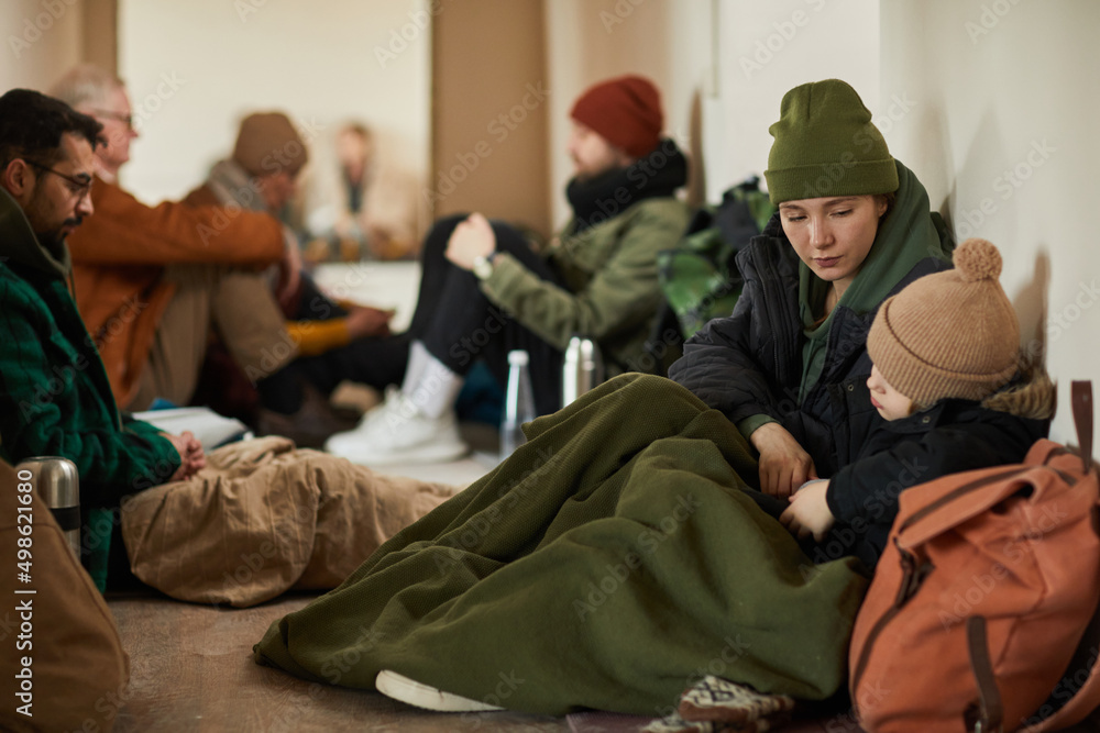 Caucasian young mother with child in refugee shelter sitting on floor and covered with blankets trying to keep warm - obrazy, fototapety, plakaty 