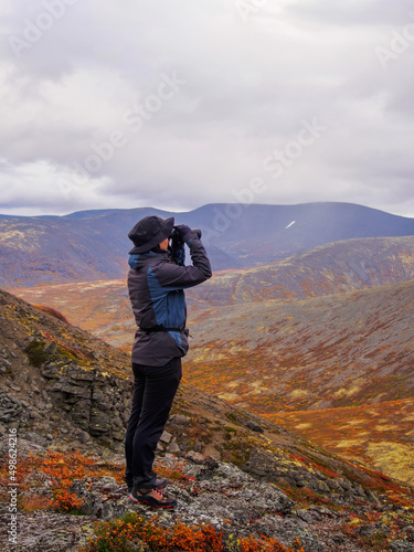 A woman in a hat on a mountain slope looks through binoculars in autumn against the backdrop of a lake and mountains. view from the back.