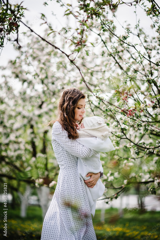 Mom hug baby girl, daughter in beautiful dresses walking on the green lawn in blooming apple blossom garden. Happy family concept. Mom hug baby girl, daughter in beautiful dresses walking on the green lawn in blooming apple blossom garden. Happy family concept.