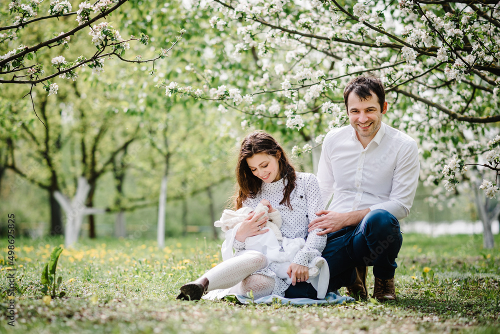Obraz premium Young beautiful family with baby child hugging each other in blossoming apple garden. Happy parents spending time together, outside, on vacation, outdoors. The concept of family holiday.