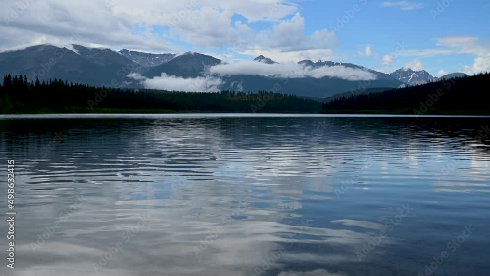 Calm mountain lake reflects mountains, trees and clouds. Small waves create patterns in the water.
Calm mountain lake reflects mountains, trees and clouds. Small waves create patterns in the water.