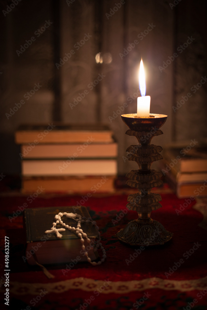 Still life with rosary on the prayer book, candle in vintage old candlestick and books near the window. Vintage old tablecloth on the table. Christianity concept. Time of prayer. Still life with rosary on the prayer book, candle in vintage old candlestick and books near the window. Vintage old tablecloth on the table. Christianity concept. Time of prayer.