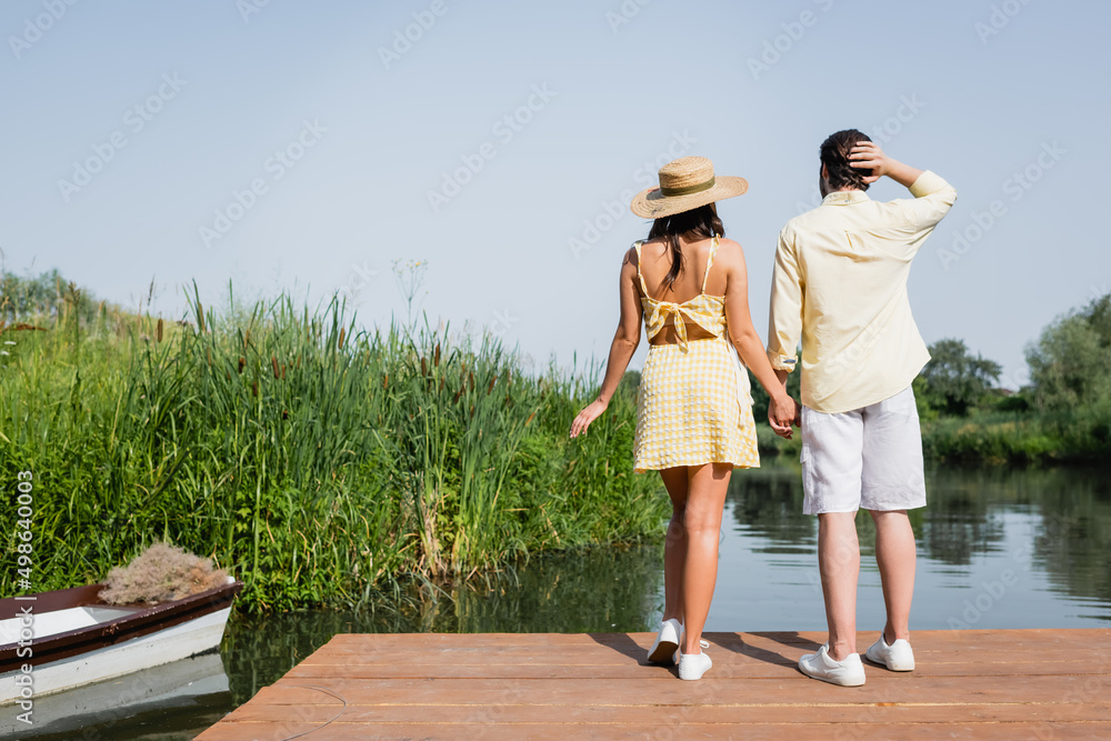 back view of young couple in summer clothes holding hands and standing on pier near lake.