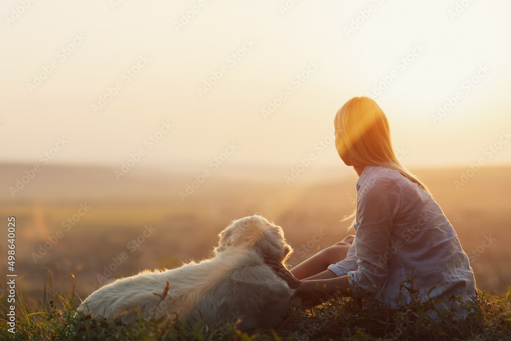 Blonde woman sitting at mountain top with white dog and looks at the valley at sunset.