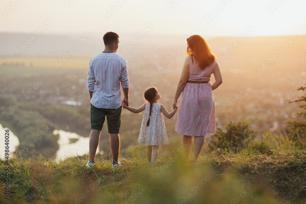 Family and childhood. Little daughter holding hands of dad and mom on the hill on background of sunset. Walking with a small kid in nature.