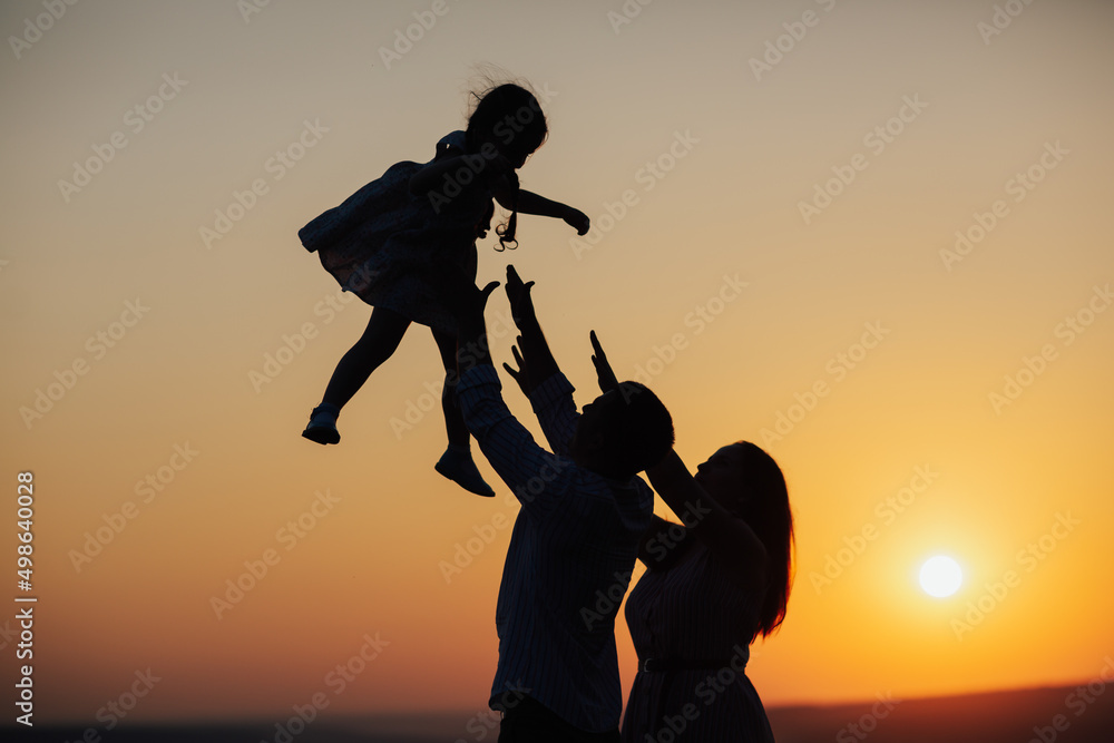 Silhouette of father and mother playing with toddler daughter at sunset. Happiness moments with family, concept of vacation, time for family.