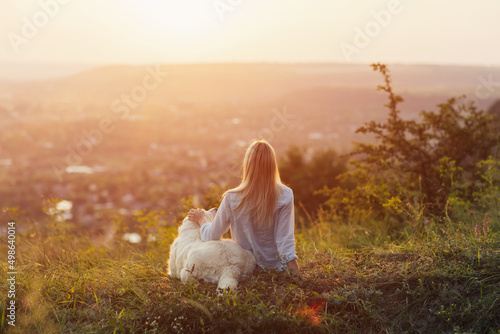 Girl with her golden retriever dog sitting on hill during summer sunset watching a beautiful landscape.