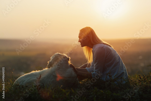 Happy pretty girl sitting on a lawn with a golden retriever dog at sunset. People with pets outdoors. © eduard