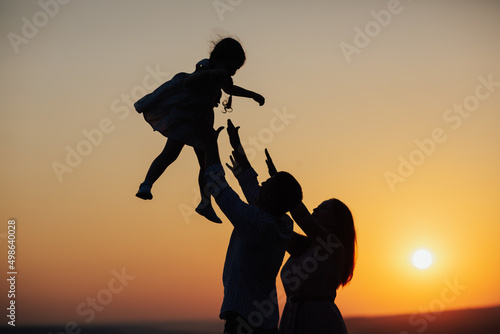 Obraz na płótnie Silhouette of father and mother playing with toddler daughter at sunset. Happiness moments with family, concept of vacation, time for family. w salonie