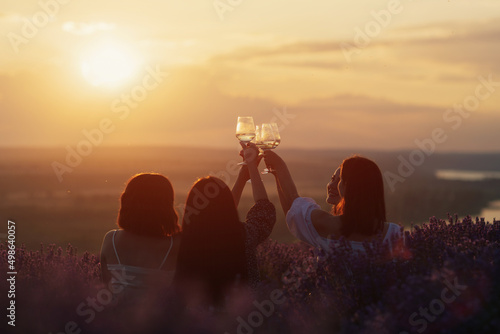 Young women having nice picnic in the lavender field, cheerful talk, clink glasses with wine on bright summer sunset. Cheers. Concept of party, joy, fun and friendship.