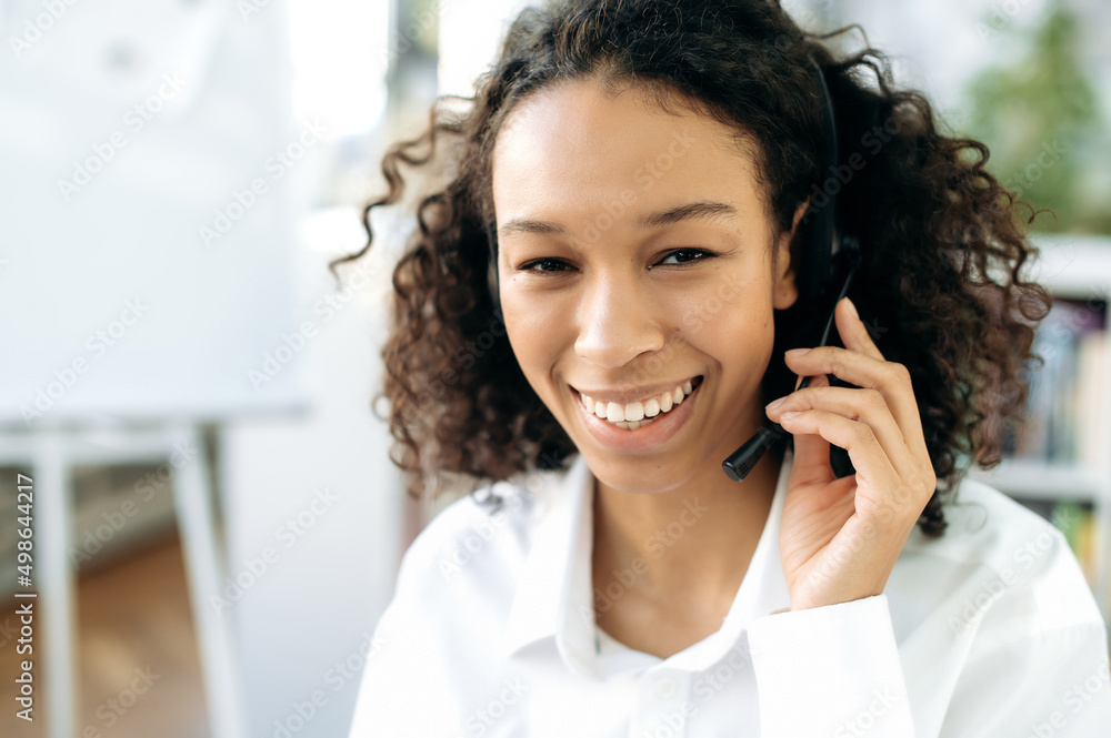 Portrait of a charming, friendly African American curly haired girl with headphones, call center operator, hotline worker, online consultant, wearing white shirt, looking at the camera, smiling Portrait of a charming, friendly African American curly haired girl with headphones, call center operator, hotline worker, online consultant, wearing white shirt, looking at the camera, smiling