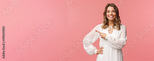 Pleasant good-looking smiling blond woman with blond short wavy hair, white tender dress, looking camera and pointing finger left to show you best price, good offer, promo concept, pink background