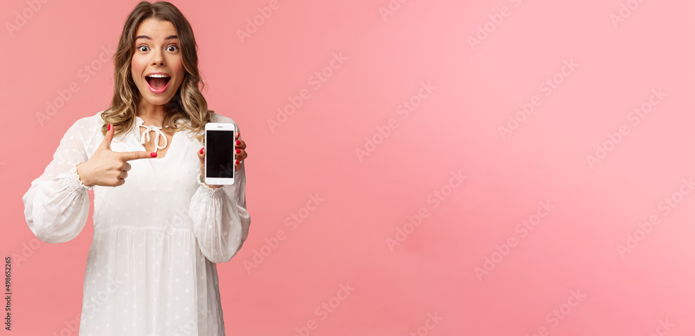 Portrait of impressed, excited young blond woman showing something awesome on display, pointing mobile phone screen and smiling astonished, brag with her recent match on dating app - obrazy, fototapety, plakaty 