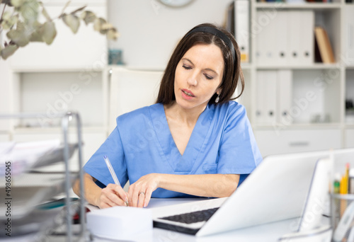 Skillful woman physician sitting at table in her office and writing.