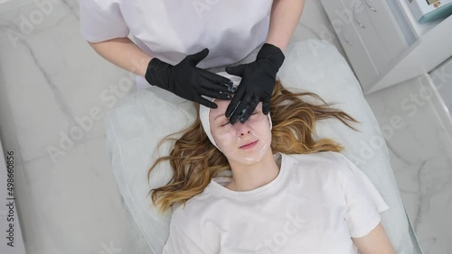 Cosmetologist applies a woman a therapeutic mask on her face. Woman in a spa salon on cosmetic procedures for facial care. Beautician makes a massage of the face with a moisturizing mask on a face.