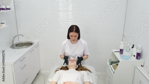 Cosmetologist applies a woman a therapeutic mask on her face. Woman in a spa salon on cosmetic procedures for facial care. Beautician makes a massage of the face with a moisturizing mask on a face.