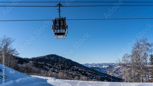 A funicular cab with attached skis moves along ropes over a snow-covered valley. Footprints in the snow. A row of bright flags on the edge of the plateau. Mountains against the sky. Altai. Manzherok