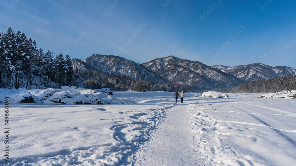 Two people are walking along a path trodden in the snow. Picturesque snow-covered boulders and coniferous trees are visible on the sides. A mountain range against a blue sky. A sunny winter day. Altai