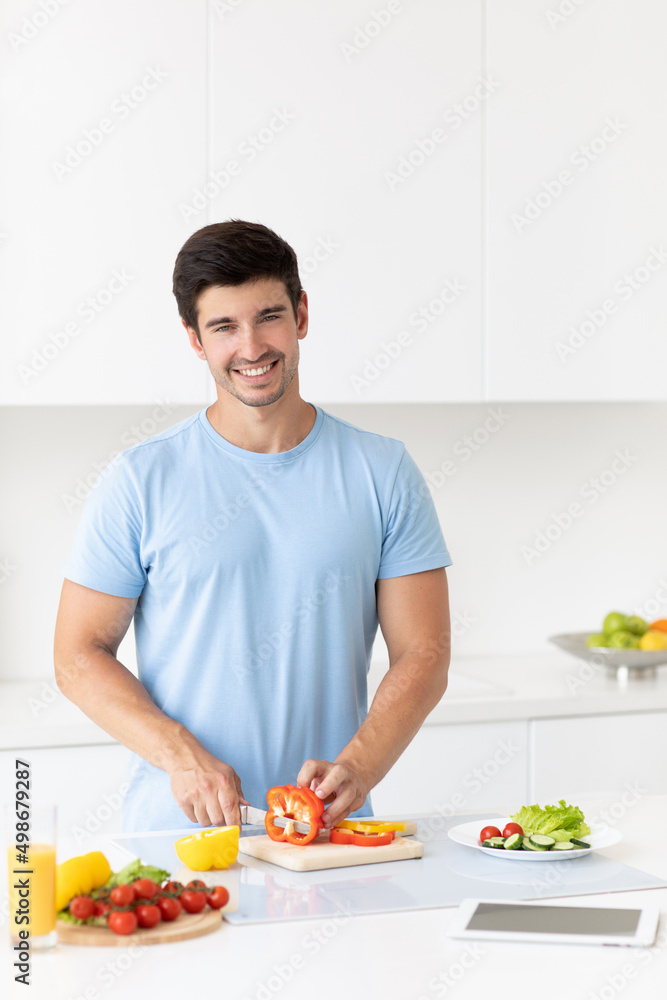 Handsome young man in a blue t-shirt prepares a vegetable salad in a bright kitchen Handsome young man in a blue t-shirt prepares a vegetable salad in a bright kitchen