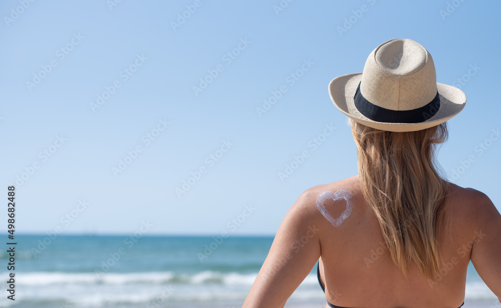 Young woman in hat  sits on beach. On back drawn heart by sunscreen.  Vacation, travel and holiday on sea in summer. Protect your skin by cream or lotion