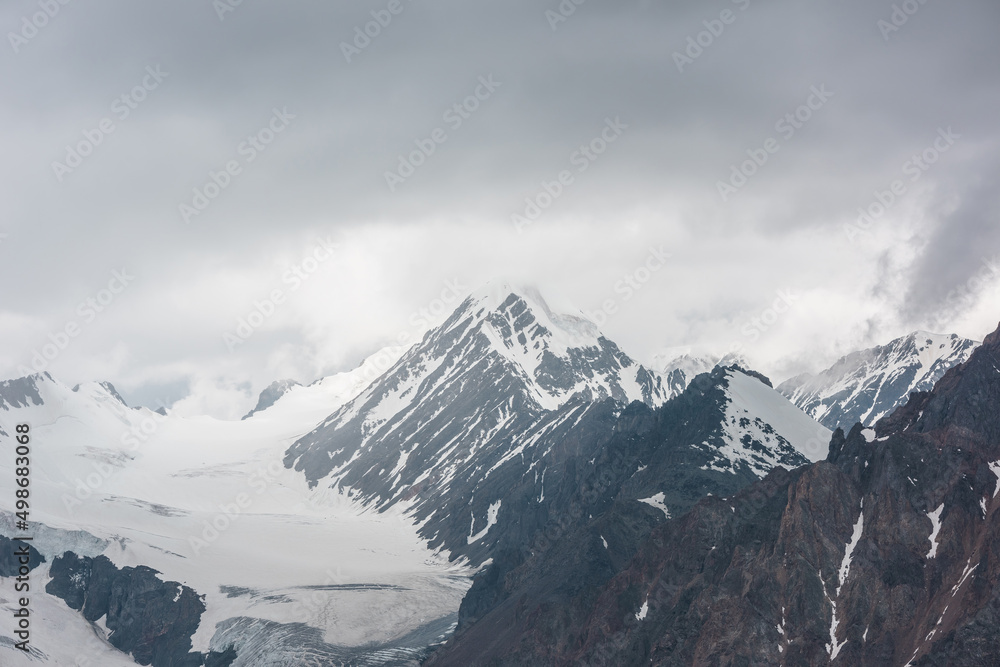 Atmospheric landscape with sharp rocks and high snowy mountain top in rainy low clouds at overcast. Dramatic gloomy scenery with large snow mountains and glacier in gray cloudy sky at rainy weather.