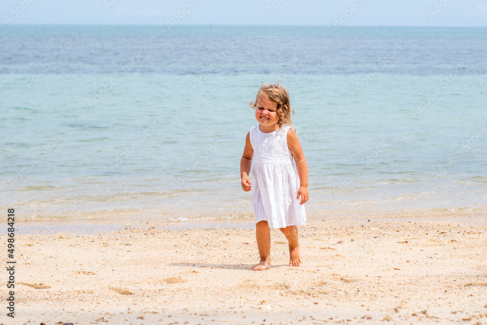 A little girl with curly hair in a white dress walks along a sandy beach on the shore of the sea, ocean and smiles. Sea holidays, travel and beach holidays with children