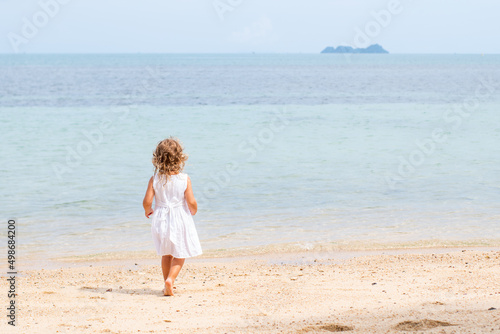 A little girl with curly hair in a white dress walks along a sandy beach on the shore of the sea  ocean and smiles. Sea holidays  travel and beach holidays with children