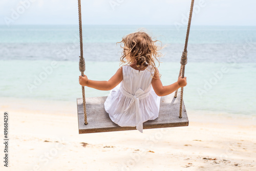 A little girl with curly hair in a white dress is swinging on a swing on a sandy beach by the sea, ocean. Sea holidays, travel and beach holidays with children